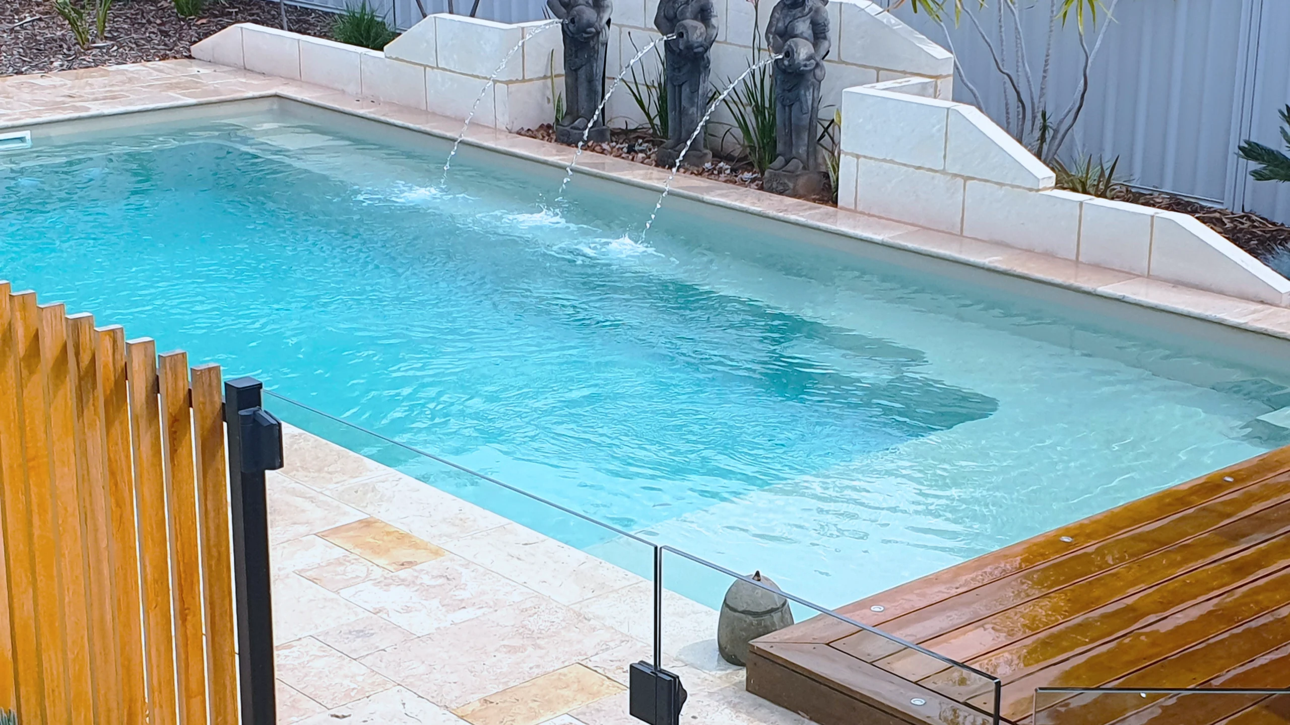 The Reflection with Splash Deck 8.50m fibreglass swimming pool in Diamond Sand is installed in a landscaped backyard in Barmera, South Australia. The pool is surrounded by light beige stone paving, warm timber decking, decorative water features and lush greenery. The Diamond Sand finish creates a soft blue-green water colour that complements the natural tones of the paving and timber, creating a tranquil and inviting outdoor retreat.