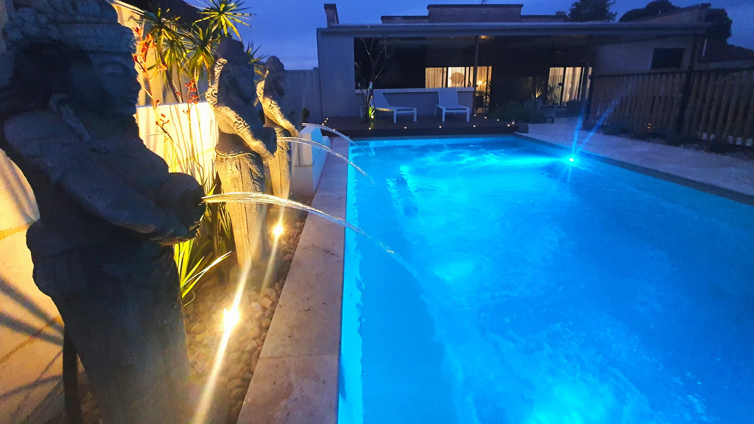 The Reflection with Splash Deck 8.50m fibreglass swimming pool in Diamond Sand is installed in a landscaped backyard in Barmera, South Australia. The pool is surrounded by light beige stone paving, warm timber decking, decorative water features and lush greenery. The Diamond Sand finish creates a soft blue-green water colour that complements the natural tones of the paving and timber, creating a tranquil and inviting outdoor retreat.