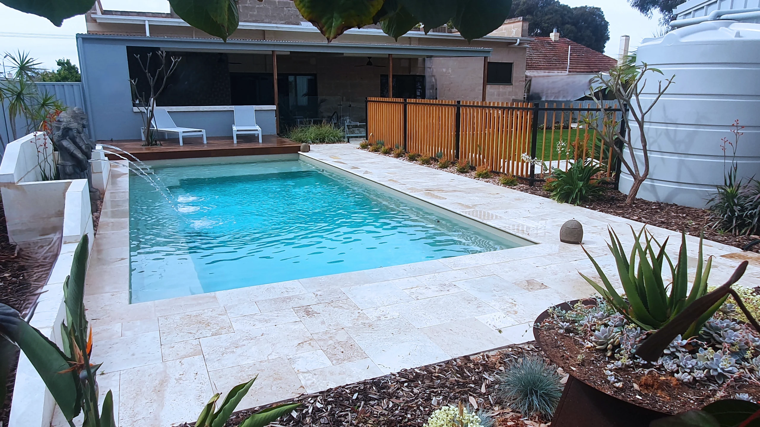 The Reflection with Splash Deck 8.50m fibreglass swimming pool in Diamond Sand is installed in a landscaped backyard in Barmera, South Australia. The pool is surrounded by light beige stone paving, warm timber decking, decorative water features and lush greenery. The Diamond Sand finish creates a soft blue-green water colour that complements the natural tones of the paving and timber, creating a tranquil and inviting outdoor retreat.